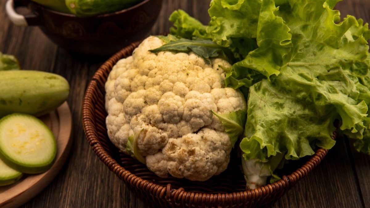 A whole cauliflower and a head of green lettuce in a wicker basket, with courgettes and cucumbers on a wooden table