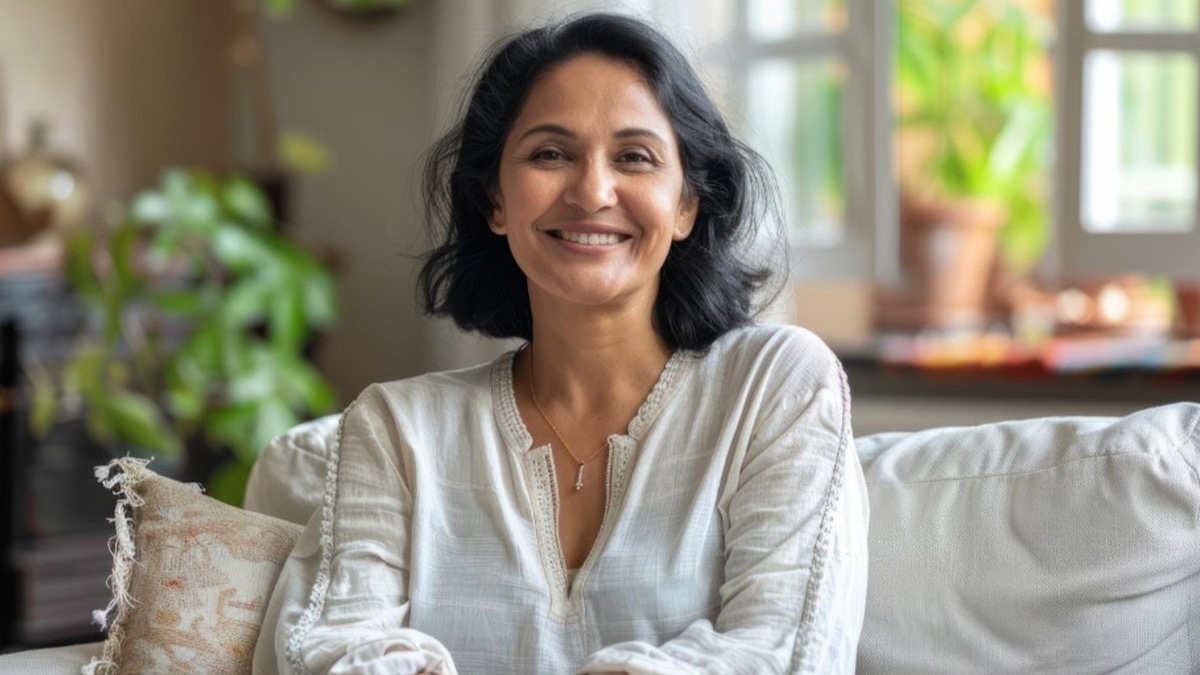 सफ़ेद कढ़ाई वाले कुर्ते में घर पर हल्के रंग के sofa पर बैठी मुस्कुराती हुई एक अधेड़ उम्र की भारतीय महिला