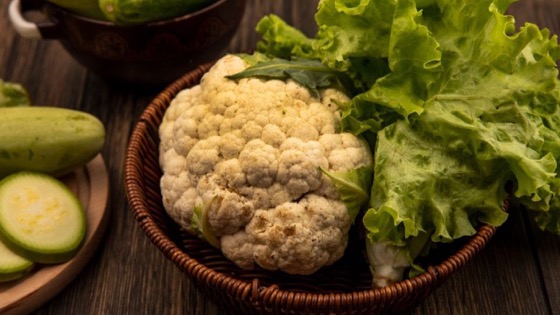 लकड़ी की मेज़ पर टोकरी में रखी एक पूरी गोभी और हरा lettuce, पास में तोरी और खीरे